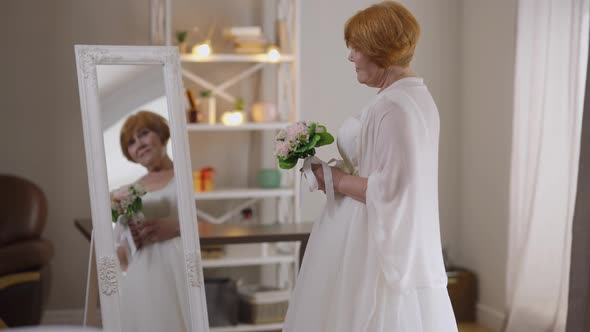 Side View Portrait of Worried Happy Senior Bride in Wedding Dress with Bridal Bouquet Admiring alt