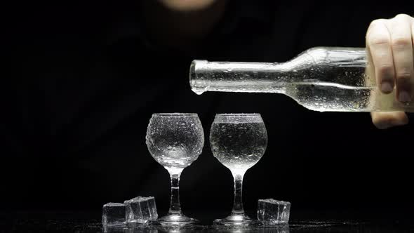 Barman Pour Frozen Vodka From Bottle Into Two Glasses with Ice. Black Background alt