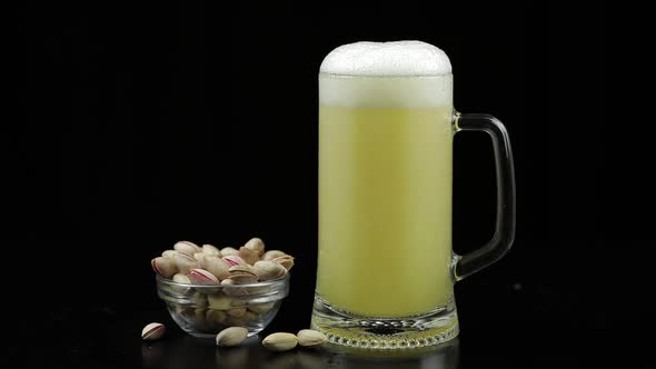 Beer Is Pouring Into Glass on Black Background. Bowl of Pistachios Nuts alt