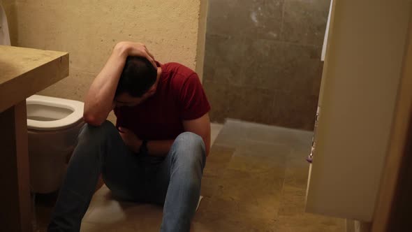 Young Sick Guy is Sitting on the Floor of Bathroom Next to Toilet and Holding His Belly alt
