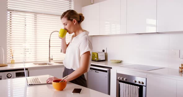 Woman using laptop while having coffee in kitchen alt