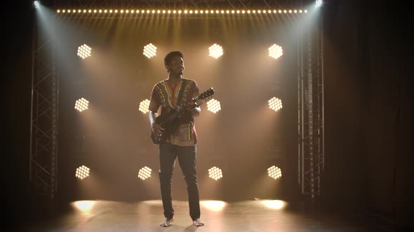 Musician in Ethnic African Costume Plays the Guitar in a Dark Studio. Black Barefoot Man Closed His alt