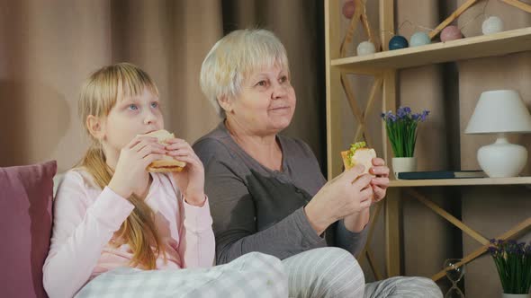 Active Elderly Woman with Granddaughter Together Watching TV and Eating Sandwiches alt