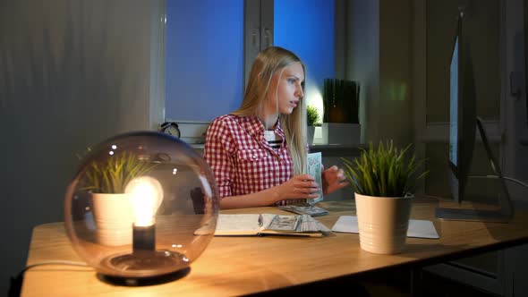 Woman Counting Cash at Working Desk. Attractive Blond Female in Checkered Shirt Sitting at Night in alt