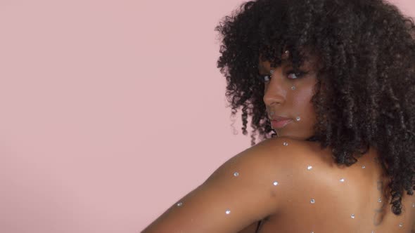 Mixed Race Black Woman with Curly Hair Covered By Crystal Makeup on Pink Background in Studio alt