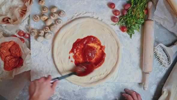 Hand is Spreading Tomato Sauce on Raw Pizza Dough with Black Ladle