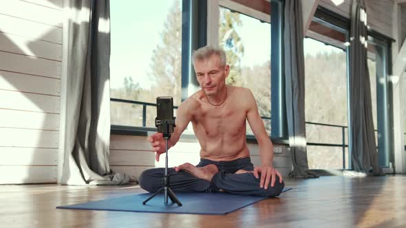 Mature Man Sitting Cross Legged on the Floor Doing Online Workout in Front of the Phone alt