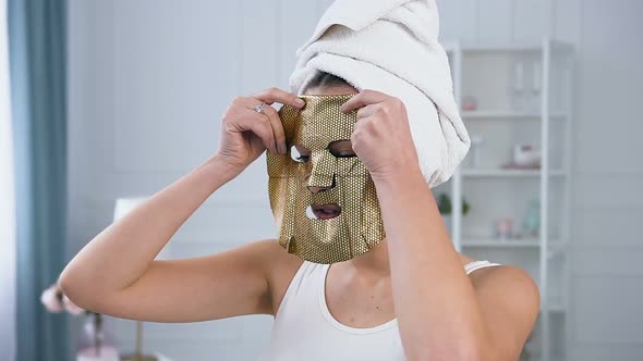 Young Caucasian Woman Applying Rejuvenating Cosmetic Golden Tissue Mask on Face. alt