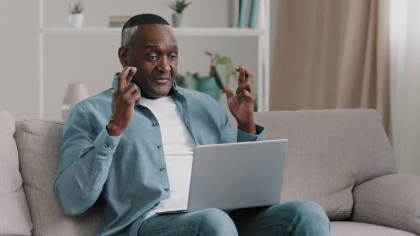 Mature African American Man Sitting at Home Looking at Laptop Screen with Hope Crossed Fingers alt