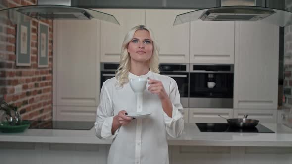 Young Blond Woman in White Shirt Enjoying Early Morning Standing on Kitchen Drinking Coffee