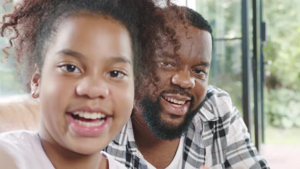 Happy African family dad and daughter having fun and using mobile phone video call on sofa at house alt