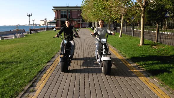 Man and Girl on Electric Scooters Are Riding Through Publick Park at Morning alt