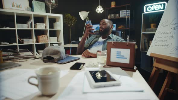Thoughtful Afro Male Professional Looking Mobile Phone in Hipster Office. alt