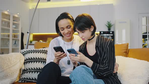 Two Young Girls Look at Their Smartphones and Have Fun Sitting on the Couch at Home alt