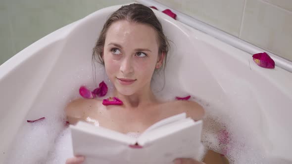 High Angle View Portrait of Charming Slim Confident Woman Lying in Bathtub with Foam Reading Book alt