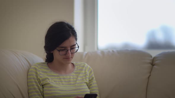 Girl Sits in Glasses on the Sofa at Home Uses the Phone and Smiling Chat with Friends