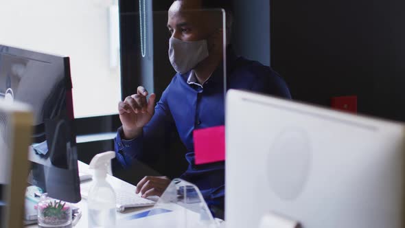 African american man wearing face mask using computer while sitting on his desk at modern office alt