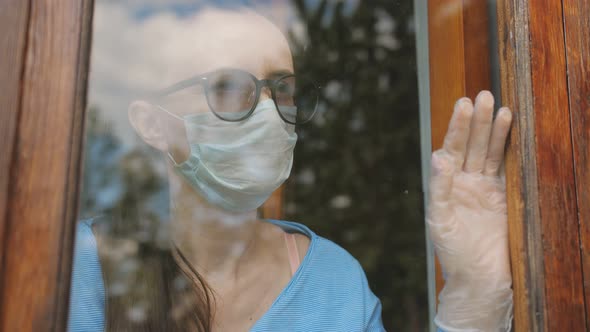 A Woman in a Medical Mask Stands Outside the Window and Looks Out Sadly
