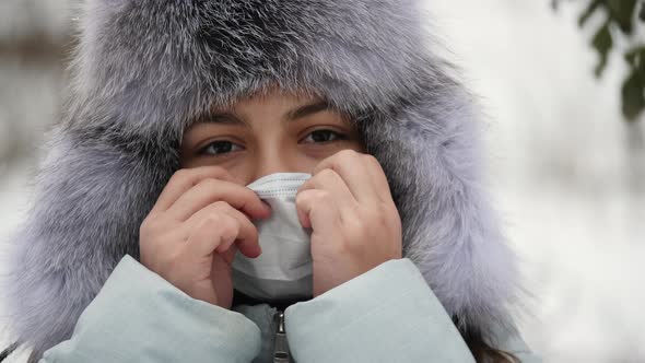 Portrait of a Teenage Girl in a Fur Hat Taking Off a Medical Mask From Her Face alt