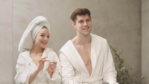 Attractive Female in Bathrobe and Towel Use Moisturizing Cream While Boyfriend Shaving Stubble