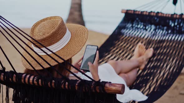 Young woman traveler lying on a hammock and using smartphone at the beach
