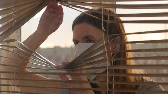 Woman in Protective Mask Opening Blinds and Looking Out the Window 