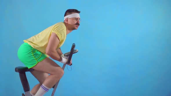 Funny Young Man From the 80's with a Mustache and Glasses on Exercise Bike on a Blue Background Slow alt