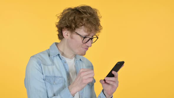 Redhead Young Man Celebrating on Smartphone Yellow Background alt