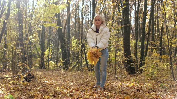 Blonde woman throws a bouquet of yellow leaves in the autumn forest. Slow motion. alt
