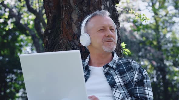 An Elderly Grayhaired Man with a Beard in Headphones Enjoys Music and Nature Sitting with a Laptop