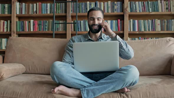 Young Man On The Couch With Laptop And Talks On Cell Phone alt
