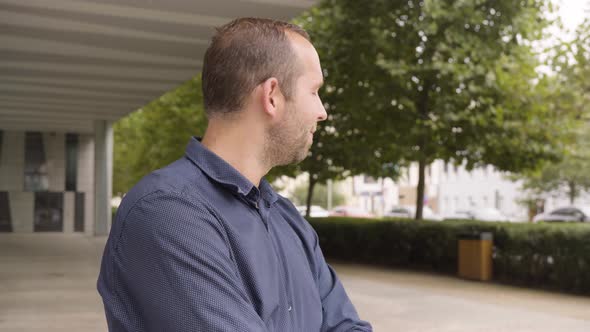 A Caucasian Man Waves at the Camera with a Smile By an Office Building in an Urban Area