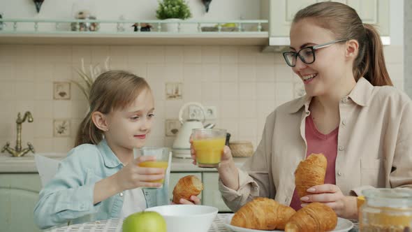 Joyful Kid and Cheerful Mom Eating Croissants and Clanging Juice Glasses in Modern House alt