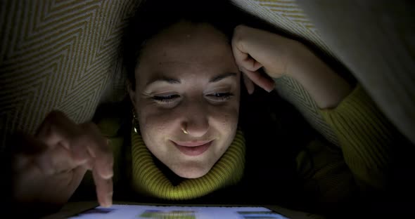 Woman on the bed under the blanket watching photos on digital tablet and smil