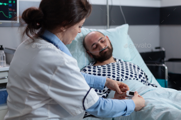 Doctor putting pulse oximeter on finger of sleeping patient with nasal cannula connected to oxygen