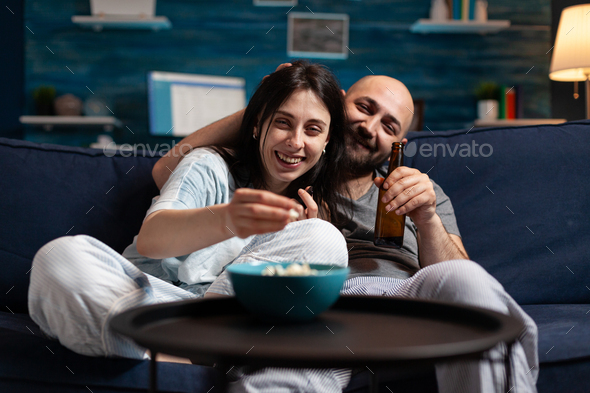 Excited couple in pajamas chilling on couch eating popcorn watching comedy movie