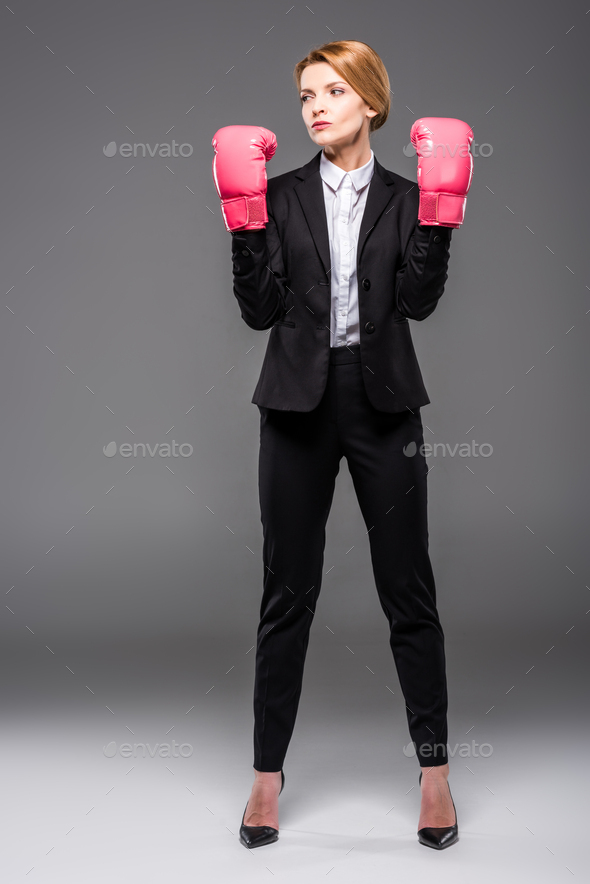 confident businesswoman posing in suit and pink boxing gloves, isolated on grey