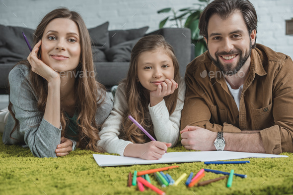 parents and daughter drawing with felt-tip pens on floor and looking at camera