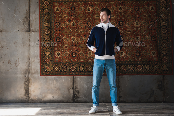 handsome young man in stylish clothes in front of rug hanging on wall