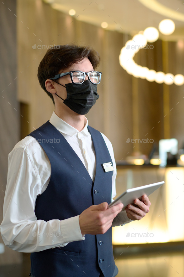 Young serious receptionist with tablet standing in lounge of modern hotel