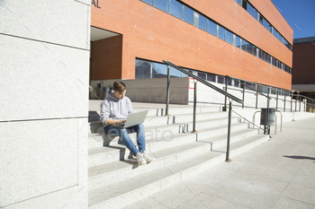 angle view young male college student sitting on stairs working on laptop computer