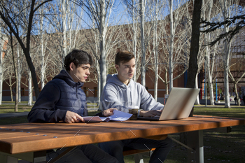 a couple of university students sitting at a table in a park with a laptop computer
