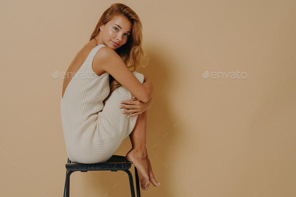 Beautiful young woman poses in profile on chair with legs curled up wears dress