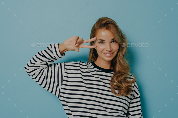 Horizontal shot of pleased lovely young woman with long hair shows peace sign over eye