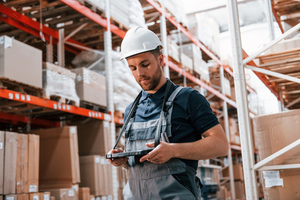 Holding tablet in hands. Employee in uniform is working in the storage at daytime - Stock Photo - Images