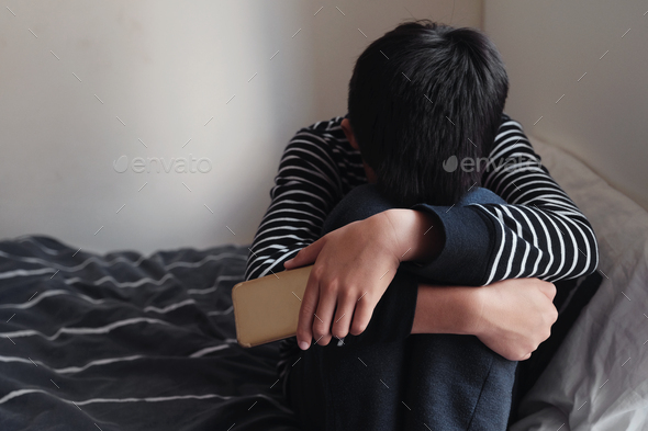 Young Asian preteen boy hugging knee in bedroom with smartphone, Cyber bullying, child mental health