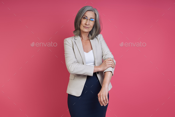 Confident senior woman rolling up sleeve on her jacket while standing against pink background