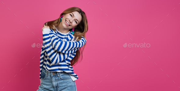 Happy young woman hugging self and smiling while standing against colored background