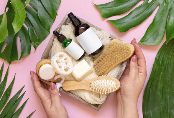 Hands holding tray of natural skin and hair care accessories near tropical leaves, mockup