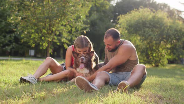 Young Couple Sitting in Park on Grass and Petting Their Dog on Sunny Summer Day